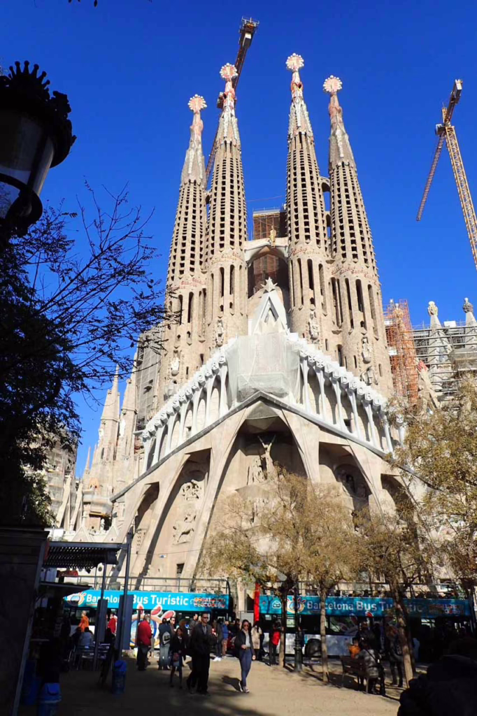 The main entrance and spires of La Sagrada Familia in Barcelona, with two construction cranes against a blue sky.